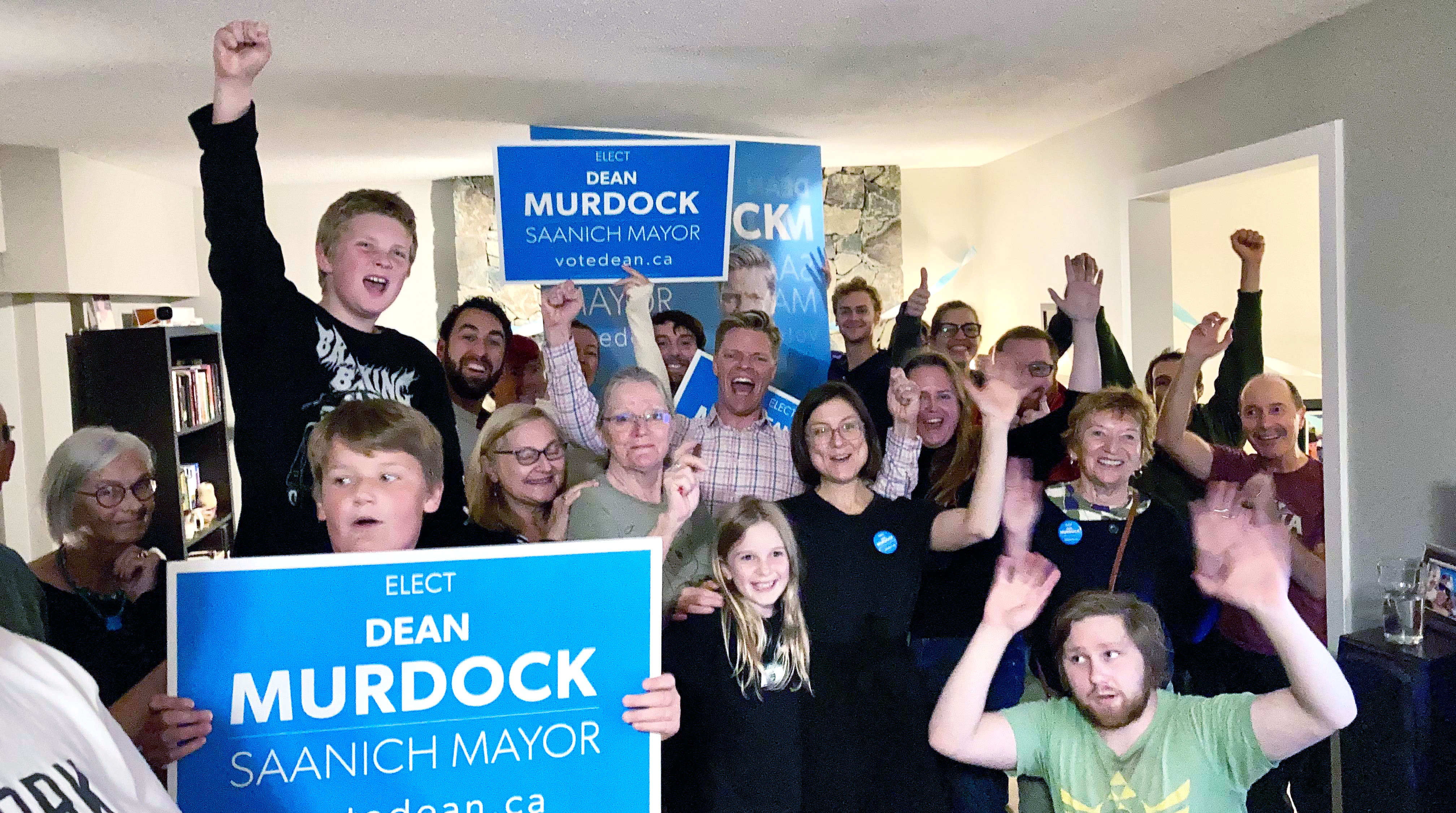 20 people cheering in a living room while holding up Dean Murdock - Saanich Mayor signs.