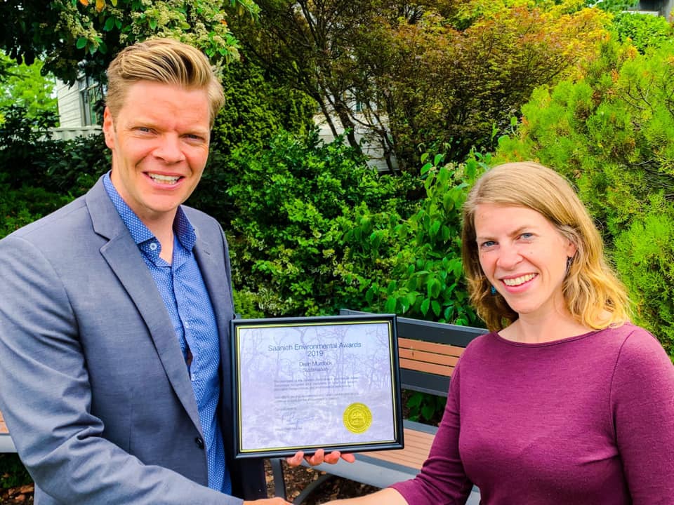 Dean holds a framed "Saanich Environmental Award" while shaking Rebecca Mersereau's hand.