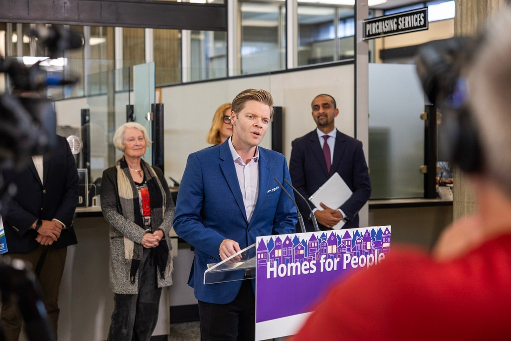 Dean speaks to the media at a glass podium with a purple sign that reads 