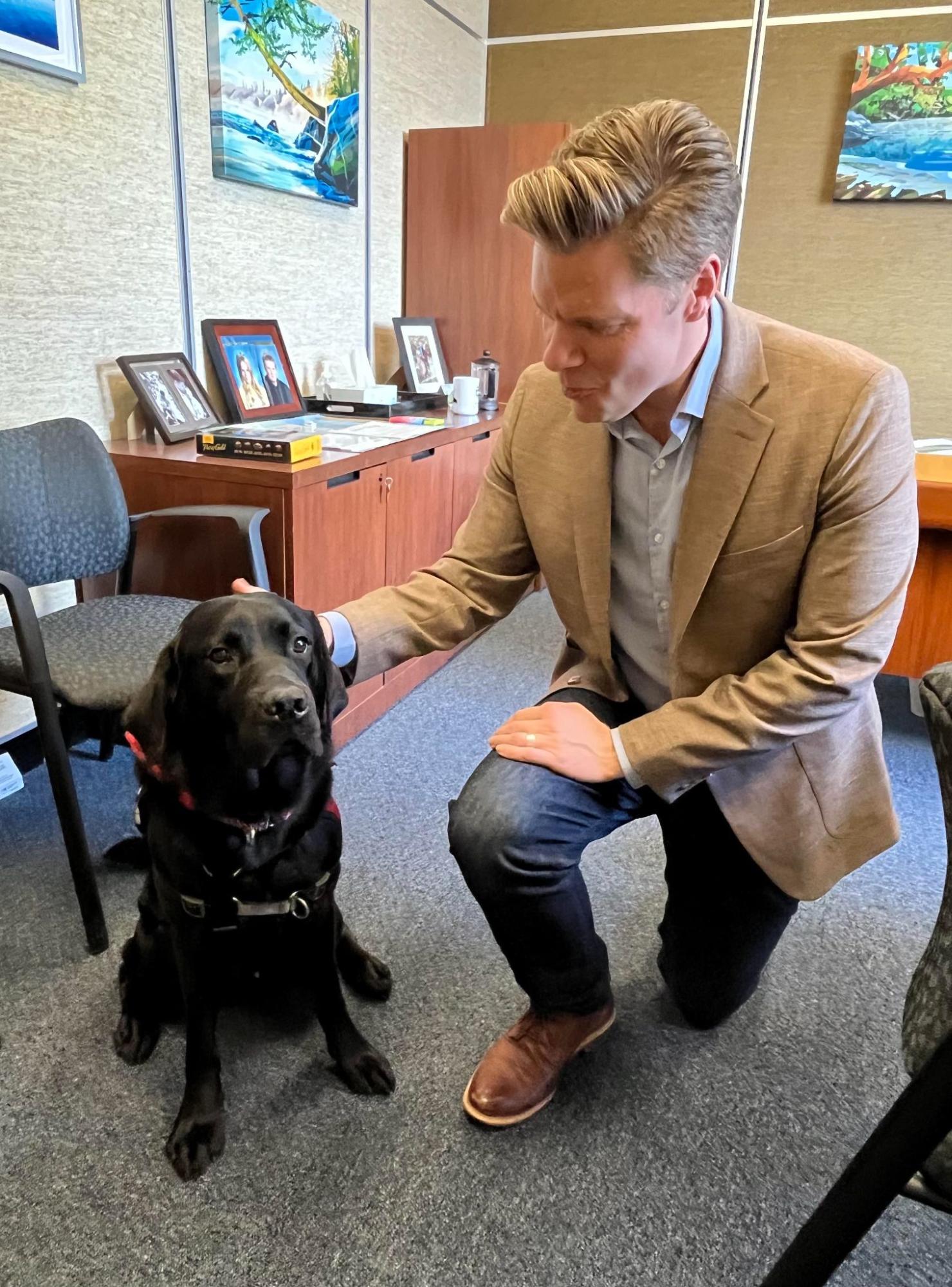 Dean kneels and pets a Black Labrador in an office