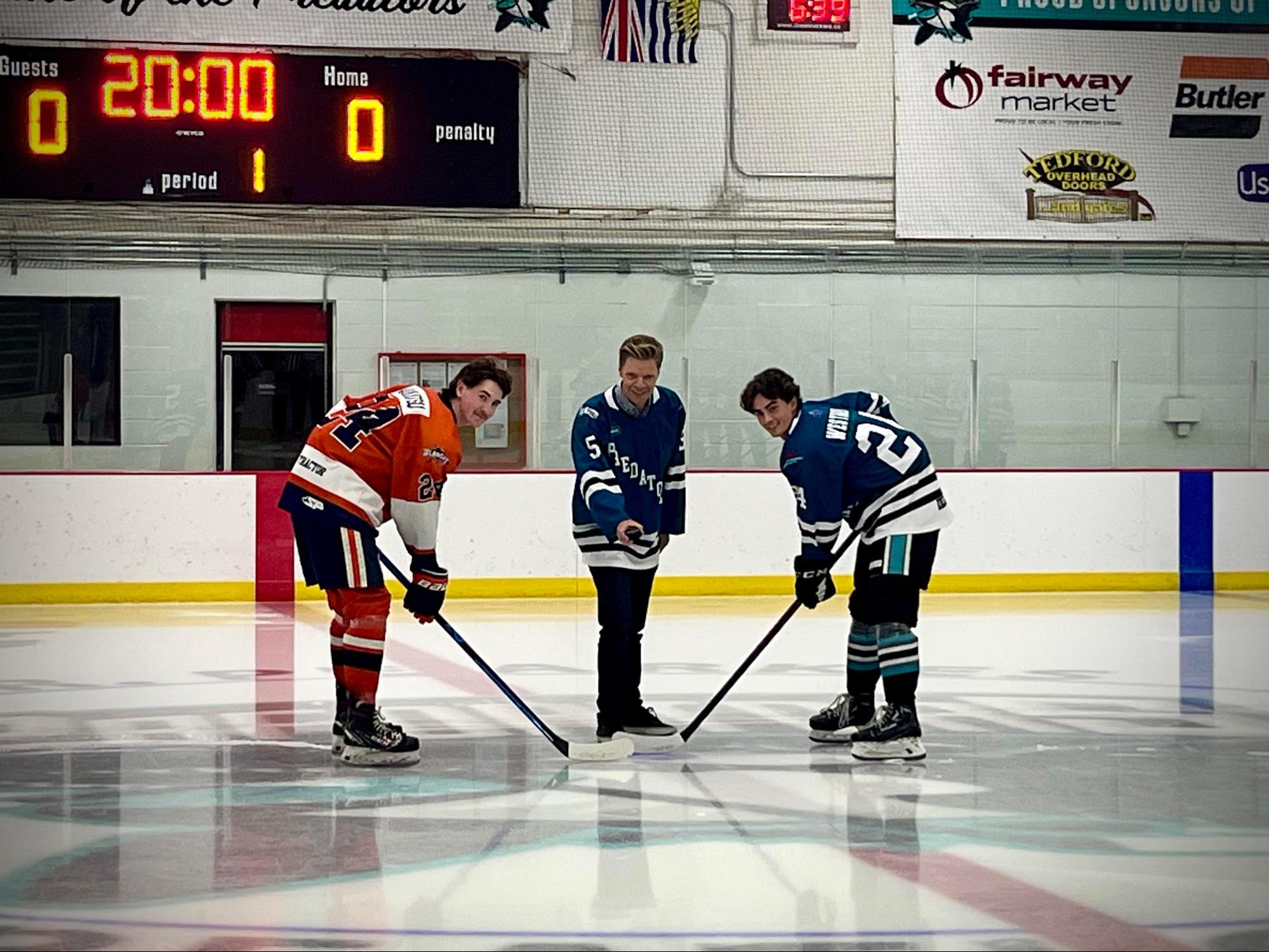 Dean stands between two hockey players on an ice rink, about to drop a puck.