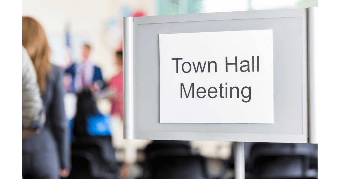 A “Town Hall Meeting” sign sits in front of a blurred man in a suit speaking to an audience